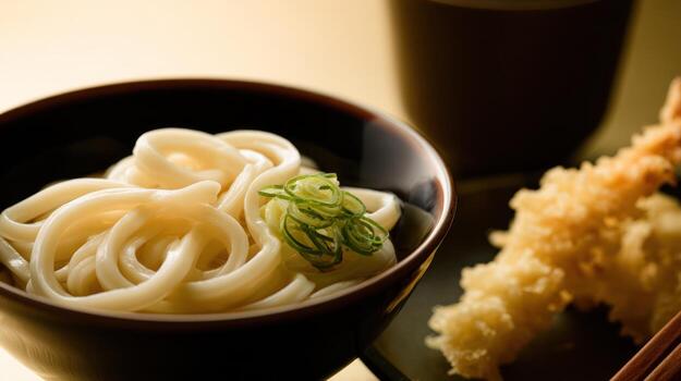 Close up of udon noodles in a bowl beside tempura and a blurred beverage photo