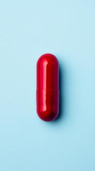 A bright red capsule rests flat against a smooth light blue backdrop, highlighting its glossy texture and simple shape photo