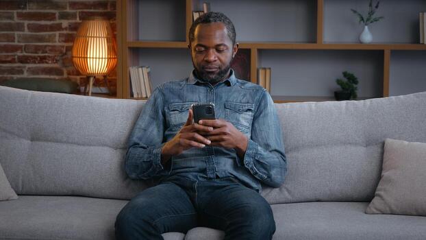 contento africano americano hombre utilizando teléfono chico masculino solo relajante en sofá con móvil teléfono inteligente a hogar disfrutando Teléfono móvil desplazamiento hojeada chateando compras en línea Internet solicitud comercio electrónico foto