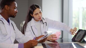 Due cardiologi impegnare nel un' dettagliato discussione su cuore malattia trattamento, utilizzando un anatomico modello e scansione nel tavoletta per illustrare loro punti mentre consulenza con un' medico stagista nel un' clinica video