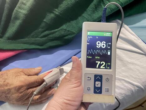 A hand holds a finger pulse oximeter displaying SpO2 96 percent and Pulse 72 BPM on a screen with waveforms. The device's sensor is attached to patient's finger, monitoring vital signs. photo