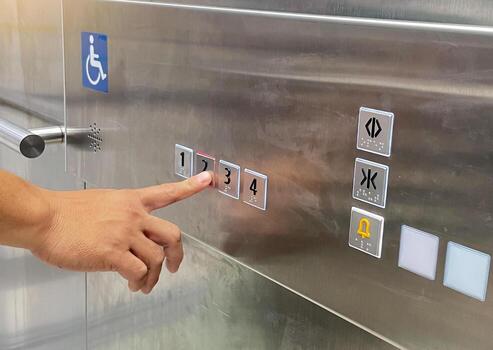 A hand presses the 2 button on a shiny metal elevator panel. The panel features floor numbers with Braille, a disabled access symbol, and other control buttons like door open-close and alarm. photo