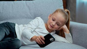 caucasian liten barn unge flicka använder sig av mobil telefon liggande på soffa på Hem med modern smartphone rullning Lycklig njuter internet social media appar dotter avkopplande på soffa leende grej missbrukare video