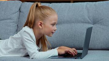caucasiano pequeno menina criança usando computador portátil deitado em sofá às casa criança filha navegando digitando computador sites em sofá jogando jogos desfrutando Internet conectados e-learning crianças tecnologia vício video
