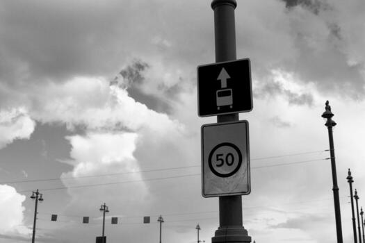 Black and white image of two road signs. One indicating bus lane and other showing speed limit of 50. Dramatic sky with clouds adds to photo atmosphere of dangerous and warning