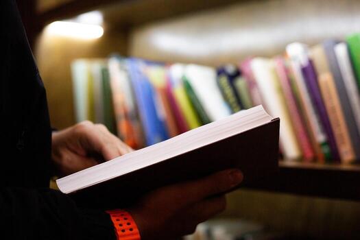 Close up of person holding book in library setting. The photo highlights moment of reading, with shelves full of books in background, creating calm intellectual atmosphere