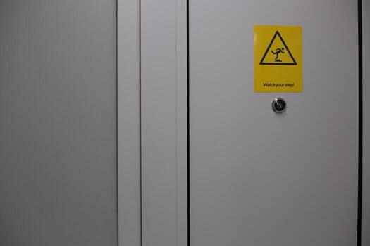 Close up image of warning sign attached to white door, with yellow triangular caution symbol and message Watch your step. The image highlights safety and caution in public spaces photo