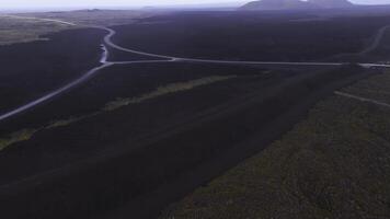 antenn se av mörk lava fält i island med korsande vägar, svag grönska, och avlägsen bergen synlig på de horisont. video