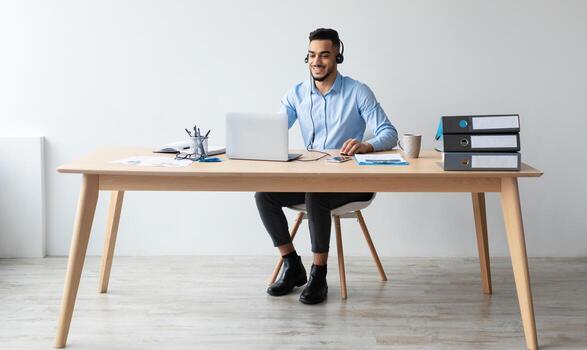 Portrait Of Smiling Middle Eastern Male Manager In Headset Working On Laptop Computer In Modern Office, Happy Guy Sitting At Desk And Consulting Clients Online Using Pc, Free Copy Space photo