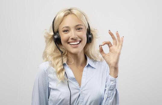Happy female call centre operator with headset showing okay gesture and smiling at camera on light studio background. Positive technical support specialist looking at camera and expressing approval photo