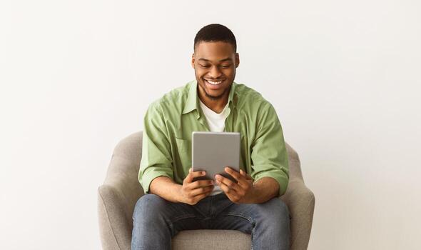 Cheerful Black Guy Using Tablet Computer Browsing Internet Or Watching Movie Online Sitting In Chair Over Gray Studio Background. Technology, Internet And Gadgets Concept photo