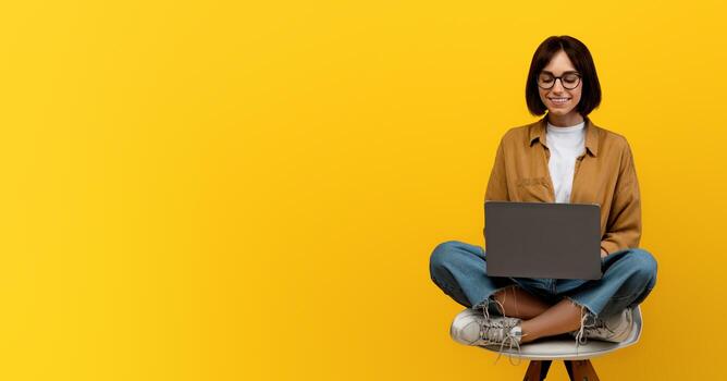 E-learning concept. Female student using laptop computer while sitting in chair over yellow studio background, copy space. Online lectures and courses, educational webinar photo