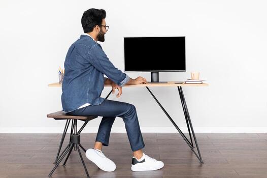 Eastern young man in casual outfit wearing eyeglasses sitting at desk, using modern pc computer with empty black display, project manager, web-designer working from home, white background, mockup photo