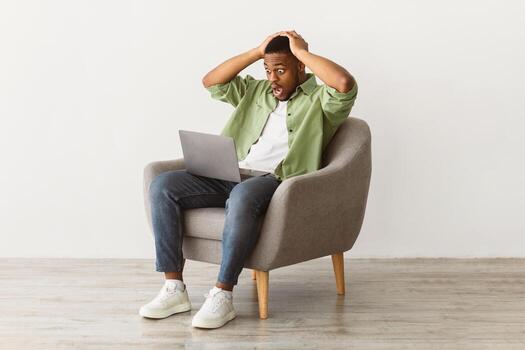 Shocked Black Man Looking At Laptop Computer Having Problem With Internet Connection Working Online Sitting In Chair On Gray Background. Oops, Negative News, Error Concept. Free Space For Text photo