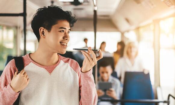 A young man smiles while using his phone's voice assistant on a bus. Other passengers are seated, some engaged with devices, creating a relaxed atmosphere during the commute. photo