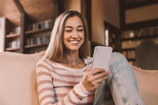 Cool Gadget And Application. Portrait of young smiling lady holding mobile phone, sending sms message, sitting on the couch in living room. Woman browsing internet, surfing web, using app, free space photo