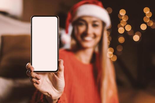 Closeup of smiling woman in red santa hat holding smartphone with white blank screen in hand, showing device, blurred background. Selective focus on gadget with empty free space for mock up photo