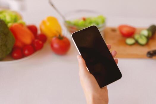 Dieting app. Woman holding cellphone with blank screen near kitchen table full of vegetables, selective focus. Healthy nutrition and weight loss mobile application. Mockup, cropped photo