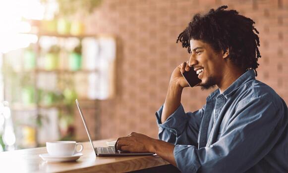 Smiling Black Freelancer Guy Talking On Cellphone While Working With Laptop At Cafe, Young African American Man Using Computer For Remote Work And Having Phone Conversation, Side View With Copy Space photo