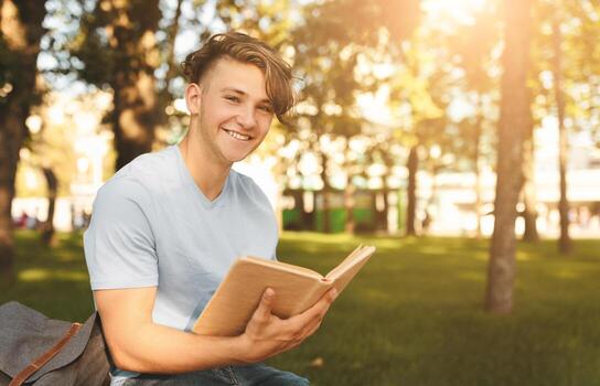 Happy student guy reading interesting book, having rest after classes, sitting on bench in parkland on sunny day and smiling at camera, free space photo