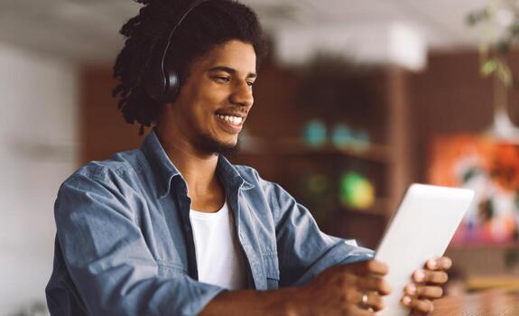 Happy Black Guy Wearing Wireless Headphones Using Digital Tablet At Cafe, Smiling Young African American Man Relaxing With Tab Computer While Sitting At Table In Modern Coffeeshop, Copy Space photo