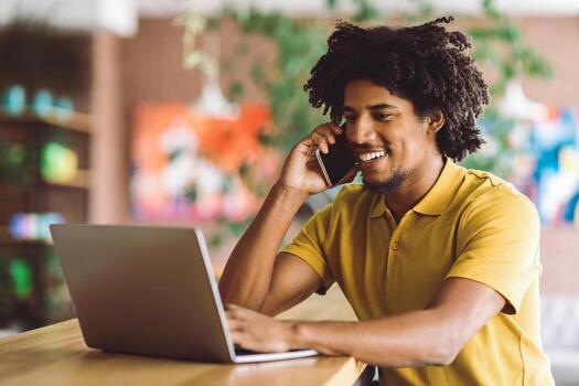 Handsome Black Young Man Talking On Cellphone And Using Laptop In Cafe, Smiling Millennial African American Guy Working Remotely On Computer And Enjoying Mobile Phone Conversation, Free Space photo