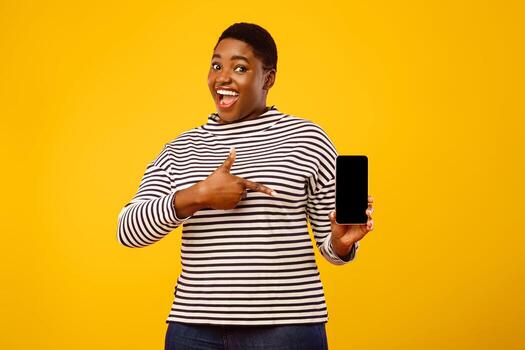 Joyful Plump African Lady Showing Cellphone With Blank Screen For Application Advertisement, Pointing Finger At Mobile Phone Standing On Pink Studio Background. Check This App. Mockup photo