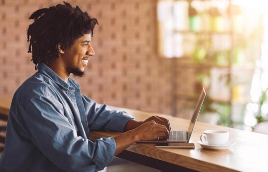 Remote Work. Young African American Freelancer Guy Working With Laptop At Modern Cafe, Millennial Black Guy Typing On Computer, Answering Emails And Drinking Coffee, Side View With Copy Space photo