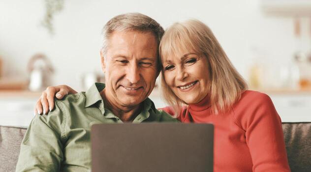 Senior Husband And Wife Using Laptop Computer Embracing While Watching Movie Onilne Sitting On Couch At Home. Older People Browsing Internet Using Gadgets. Technology Concept photo
