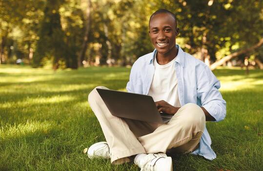 E-learning. African american student guy using laptop outdoors, having distant online lecture, sitting on grass in park. College student learning remotely, browsing internet via computer photo