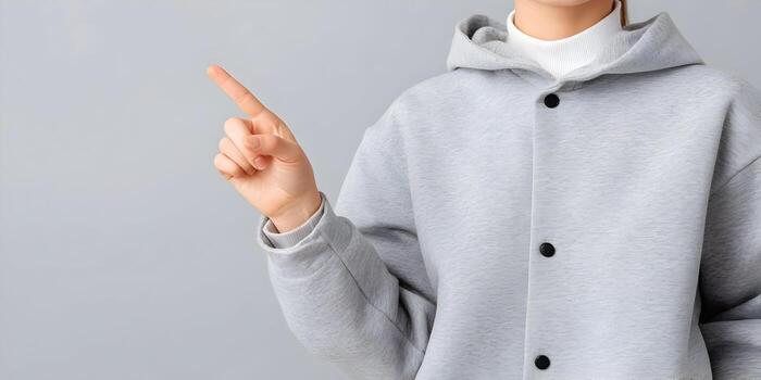 An anonymous person in a gray hooded jacket points their index finger upwards against a simple gray background photo