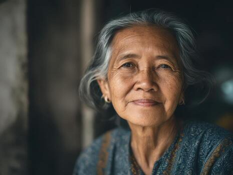 Elderly woman with gray hair and gentle smile gazes into the distance, wearing a patterned garment. Soft lighting enhances her serene expression, creating a warm, intimate atmosphere photo