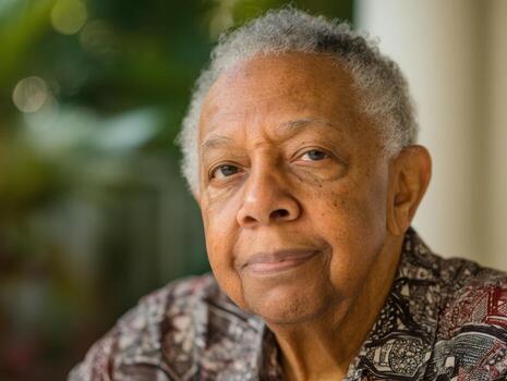 Elderly man with graying hair, wearing patterned shirt, looks directly at camera with a gentle expression. Blurred foliage in background creates a soft, natural atmosphere photo