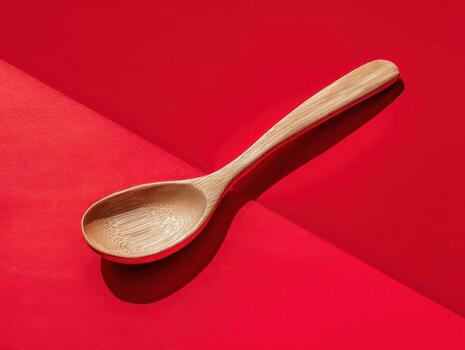 Wooden spoon rests diagonally on a smooth red surface, casting a shadow. The spoon's grain pattern is visible, and it appears to be made of a light-colored wood. No text is present photo