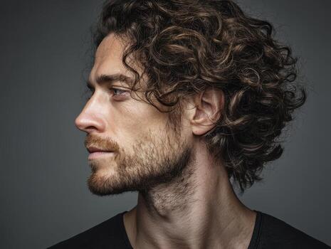 Curly-haired man with a beard and mustache looks left, displaying a strong profile. Dark-toned shirt contrasts sharply with the man's well-defined facial features and natural curls photo