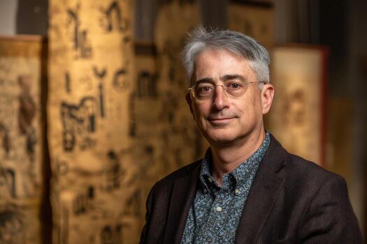 Dapper individual with gray hair and glasses, dressed in a dark blazer over a patterned shirt stands against a backdrop of rolled ancient scrolls featuring hieroglyphic-like symbols photo