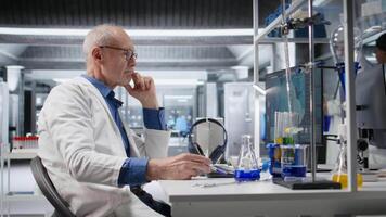 Older scientist in modern lab playing with a pen while waiting for experiment data, reflecting impatience and anxious concentration during advanced medical research in biotechnology. Camera A. video