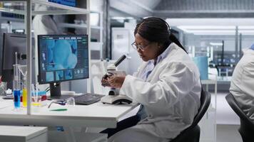 africano americano mulher analisando molecular amostras dentro uma bio laboratório, usando profissional microscópio. cuidados de saúde e bioquímica experimentos representar científico método e descoberta. Câmera b. video