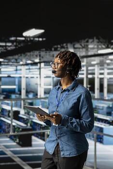 Server room admin using tablet and headset microphone to provide tech support on industrial platform in server farm. Data center IT expert resolves backend issues, coordinating remote teams. photo