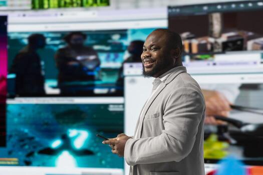 Cheerful man browses social media feed on smartphone in front of digital wall showcasing multimedia content. Happy viewer in modern space surrounded by diverse multimedia clips. photo