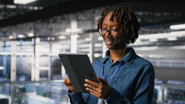 Data center engineer wearing headset microphone providing remote diagnostics using tablet. Technician monitors system health and guides users through infrastructure troubleshooting steps photo