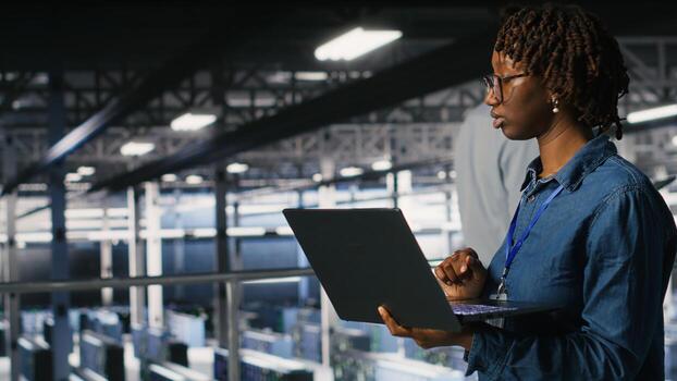 Data center computer scientist doing maintenance tasks on laptop, ensuring optimal performance. African american technician reviewing diagnostics and applying patches to server hub gear photo