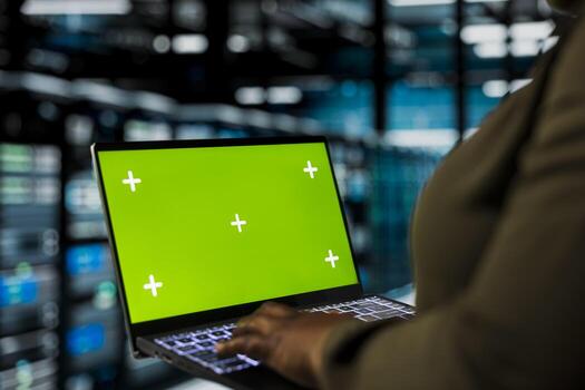 Programmer uses green screen mockup laptop to do maintenance in data center with setup helping companies stay connected. Worker walks with chroma key notebook between server hub rigs blinking lights photo