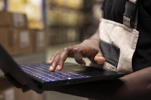 Close up of black person processing orders on laptop and checking inventory, fulfillment center operations for efficient parcel import export for e-commerce retail large business. photo