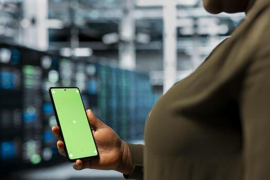 Close up of data center IT specialist using green screen smartphone to monitor mainframes network performance. African american woman in server room checks gear rigs functionality using mockup phone photo