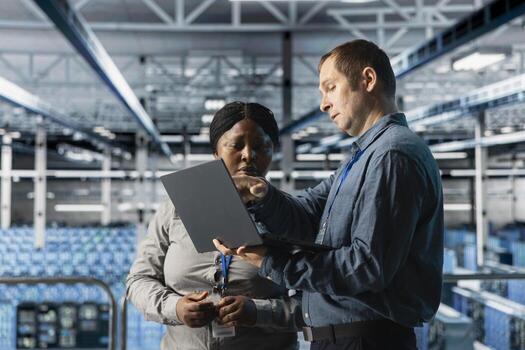 Teamworking data center technicians installing upgraded server racks systems using laptop. Server room coworkers monitoring infrastructure performance using automation tools on notebook photo