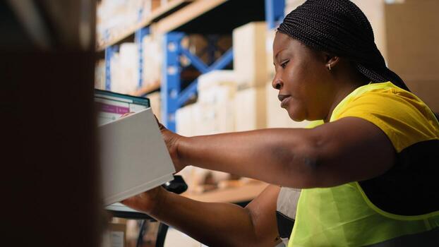 Black woman placing adhesive shipment tags on cardboard boxes, handles labeling and packaging tasks for order processing. Worker applying printed awb labels in storage depot. Camera B. photo