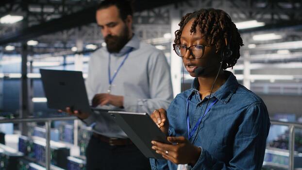 Server room admin using tablet and headset microphone to provide infrastructure tech support. Data center IT specialist resolves backend issues while coordinating with remote teams photo