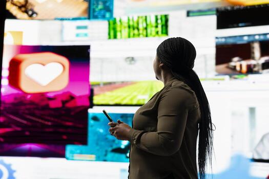 User engages with multimedia content on cellphone, sending likes across social networks. African american woman browses dashboard of streaming videos and social media feeds on digital wall photo