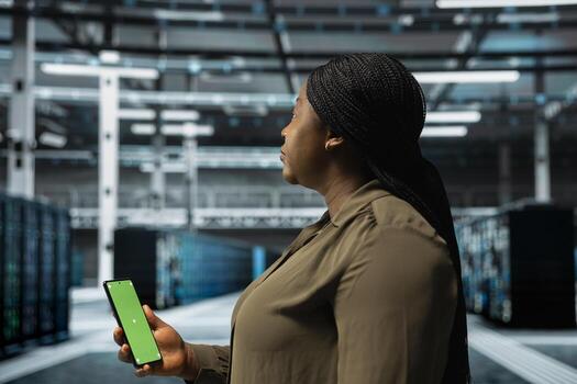Woman in data center checking mainframes performance data on mockup smartphone. African american server farm worker using green screen phone to look at system alerts, monitoring equipment photo